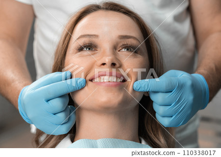 A woman sits in a dental chair, happily demonstrating her bright smile, under the sensitive and attentive gaze dentist who is ready to do everything to preserve her health and the beauty her smile. 110338017
