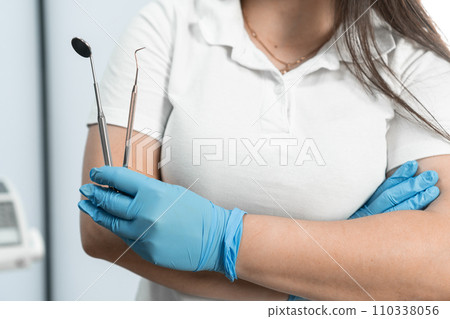 A dentist holding dental instruments in her hand, who knows how to create beautiful smiles, posing in her dental office, conveying confidence and professional expertise. 110338056