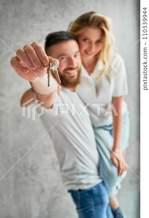 Cheerful man holding wife and smiling while showing apartment keys. Happy couple real estate house buyers demonstrating keys from new home. Focus on male hand with keys. 110339944