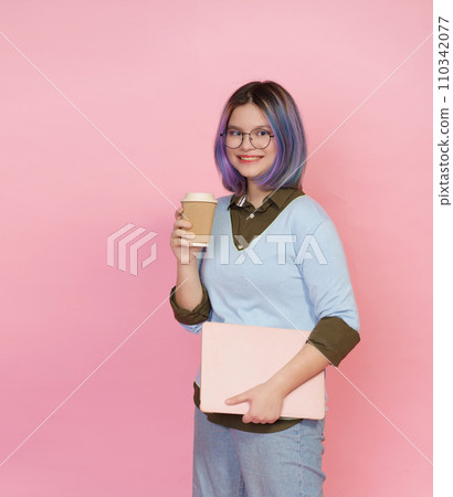Smiling pupil girl teenager, wearing glasses, immersed in studies with folded laptop and cup of coffee or tea on pink background. Student showcases joyful expression, embodying essence of modern 110342077