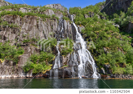 Cascading waterfall in Trollfjorden, Lofoten, bathed in sunlight amidst lush greenery Cascading waterfall in Trollfjorden, Lofoten, bathed in sunlight amidst lush greenery 110342504