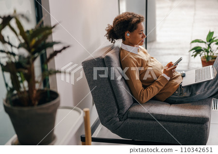 Stylish business woman is use phone while working on laptop in modern office near window 110342651