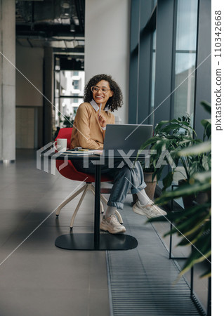 Smiling female manager in eyeglasses working on laptop while sitting in modern office near window Smiling female manager in eyeglasses working on laptop while sitting in modern office near window 110342668