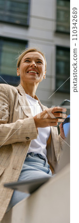 Smiling female entrepreneur use phone while working on laptop in modern office background Smiling female entrepreneur use phone while working on laptop in modern office background 110342689