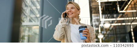 Female manager talking phone while drinking coffee standing on modern office building background 110342693