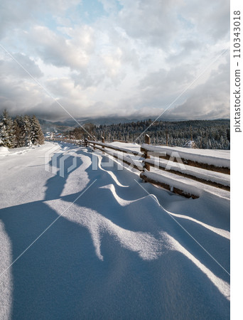 Picturesque waved shadows on snow from wood fence. Alpine mountain winter hamlet outskirts, snowy path, fir forest. High resolution image with great depth of field. 110343018