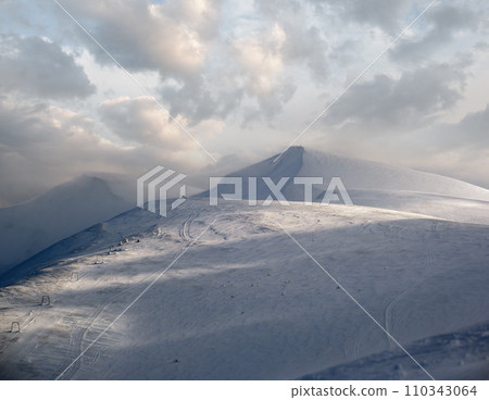 Snow covered mountain slope in last evening sunlight. Magnificent windy dusk on tops above picturesque alpine ski resort, Dragobrat, Ukraine, Carpathian Mountains. Snow covered mountain slope in last evening sunlight. Magnificent windy dusk on tops above picturesque alpine ski resort, Dragobrat, Ukraine, Carpathian Mountains. 110343064