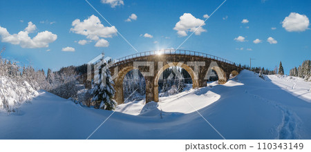 Stone viaduct (arch bridge) on railway through mountain snowy fir forest. Snow drifts on wayside and hoarfrost on trees and sunshine in sky. Stone viaduct (arch bridge) on railway through mountain snowy fir forest. Snow drifts on wayside and hoarfrost on trees and sunshine in sky. 110343149