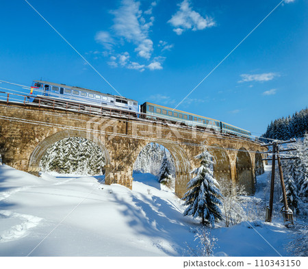 Stone viaduct (arch bridge) on railway through mountain snowy fir forest. Snow drifts  on wayside and hoarfrost on trees and electric line wires. 110343150