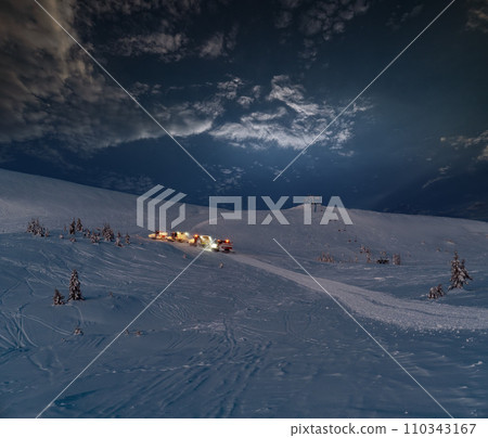 Snow groomers (snowcat ratrack machines) on night predawn excursion ride to winter mountain top with freeriders snowboarders. Alpine ski resort Dragobrat, Ukraine, Carpathian Mountains. 110343167