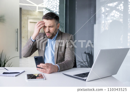 Worried young male businessman sitting in the office at the table and looking disappointedly at the phone screen holding his head. Worried young male businessman sitting in the office at the table and looking disappointedly at the phone screen holding his head. 110343525