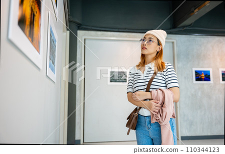 Asian woman standing she looking art gallery in front of colorful framed paintings pictures on white wall, people watch at photo frame to leaning against at show exhibition artwork gallery, Side view 110344123