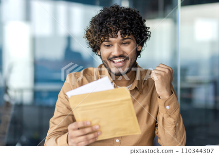 Businessman received postal envelope notification letter with good news, man celebrates holding hand up, joyfully reading news, sitting inside office with laptop. 110344307