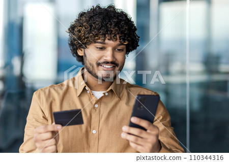 Businessman inside office with telephone and bank credit debit card in hands, man uses application on smartphone to buy in online store, satisfied worker inside office with laptop. 110344316