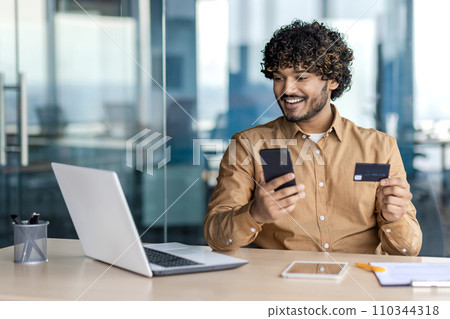 Businessman inside office with telephone and bank credit debit card in hands, man uses application on smartphone to buy in online store, satisfied worker inside office with laptop. Businessman inside office with telephone and bank credit debit card in hands, man uses application on smartphone to buy in online store, satisfied worker inside office with laptop. 110344318