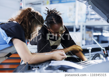 Team of women in auto repair shop working together on fixing car, discussing best options. Teamworking mechanics collaborating on servicing broken vehicle, checking for damaged fuel tank Team of women in auto repair shop working together on fixing car, discussing best options. Teamworking mechanics collaborating on servicing broken vehicle, checking for damaged fuel tank 110345315