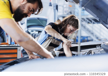 Repairman and coworker collaborating on servicing broken vehicle, checking for faulty alternator. Experienced mechanics in auto repair shop working together on fixing car, discussing best options 110345329