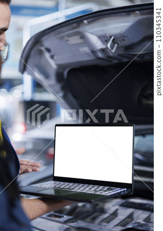 Experienced serviceman in garage using mockup laptop to follow checklist while doing maintenance on car. Hardworking professional in repair shop does checkup on vehicle with isolated screen device 110345341