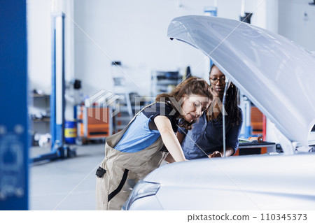 Knowledgeable mechanic helping client with car checkup in auto repair shop. Employee in garage facility looking over automobile parts with woman, mending her vehicle ignition system during inspection Knowledgeable mechanic helping client with car checkup in auto repair shop. Employee in garage facility looking over automobile parts with woman, mending her vehicle ignition system during inspection 110345373