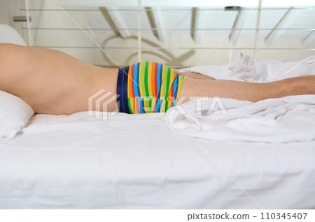Close-up gay man of non traditional sexual orientation sleeping on top of white bed sheets in rainbow boxers 110345407