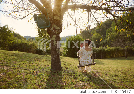 Happy young mother holding and kissing her newborn baby, swinging slowly together on the swing in the garden at sunset Happy young mother holding and kissing her newborn baby, swinging slowly together on the swing in the garden at sunset 110345437