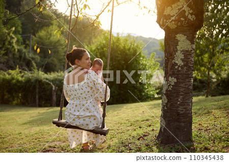 Rear view of a young adult mother holding her newborn baby while sitting on the swing in the park at sunset Rear view of a young adult mother holding her newborn baby while sitting on the swing in the park at sunset 110345438