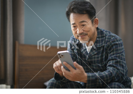 Middle-aged man in pajamas sitting on bed and looking at smartphone screen 110345886