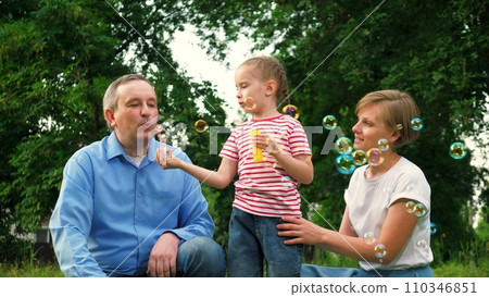 Father with mother looks at child blowing bubbles in fresh air during holidays Father with mother looks at child blowing bubbles in fresh air during holidays 110346851
