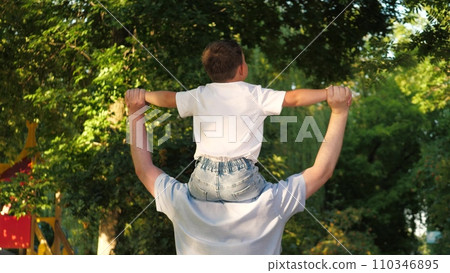 Happy father holds small pleased son on shoulders walking in city park 110346895