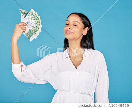 .Money, wealth and rich woman holding fan of cash and cooling herself, ready to blow or spend it all. Female lottery winner embracing luck, success and win in the studio on a blue background. 110349429