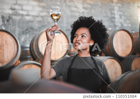 Black woman tasting wine at a winery, looking and checking the color and quality of the years produce. Young African American sommelier proud of the new addition, analyzing white wine in a cellar 110351084