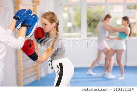 Concentrated teen girl in boxing gloves practicing punches on mitts in hands of instructor 110351124