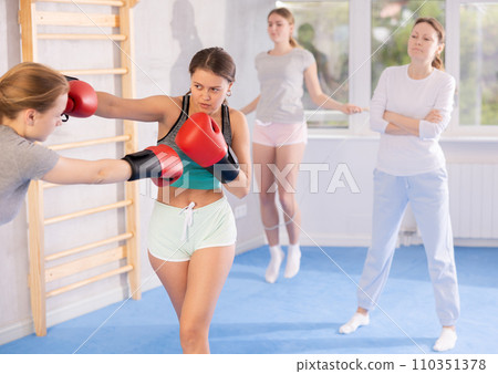 Two teenage girls in sportswear are engaged in boxing sparring in the gym under guidance of trainer in gym Two teenage girls in sportswear are engaged in boxing sparring in the gym under guidance of trainer in gym 110351378
