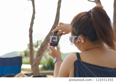 Woman wearing dark glasses smiling and taking a photo with smartphone in the park. Woman wearing dark glasses smiling and taking a photo with smartphone in the park. 110351393