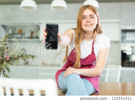 Happy teen girl in red polka dot apron showing modern smartphone with black screen in modern kitchen Happy teen girl in red polka dot apron showing modern smartphone with black screen in modern kitchen 110351411