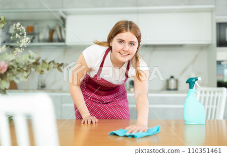 in kitchen,positive woman hostess in carved apron holds rag in hands and cleans surface of table in kitchen,positive woman hostess in carved apron holds rag in hands and cleans surface of table 110351461