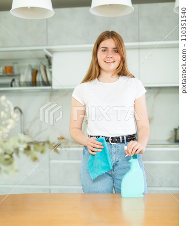 in kitchen, woman hostess in casual clothes stands and holds rag and cleaning spray in hands. 110351540