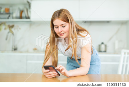 bored young woman stands in kitchen leans on countertop and surfs internet.Home pastime bored young woman stands in kitchen leans on countertop and surfs internet.Home pastime 110351580