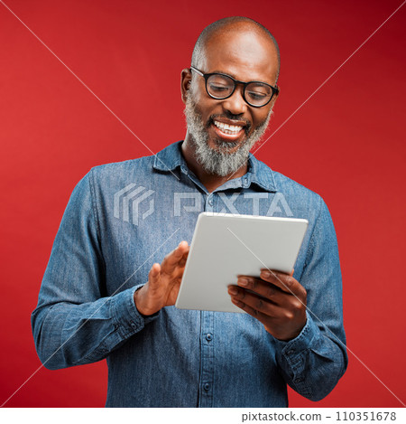 Smiling man browsing on a tablet online, networking on the internet and scrolling on an app while standing against a red studio background. Happy, mature and African male reading an email and typing 110351678