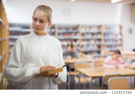 Intelligent teenager girl standing alone near bookcase in library browsing textbooks 110351784