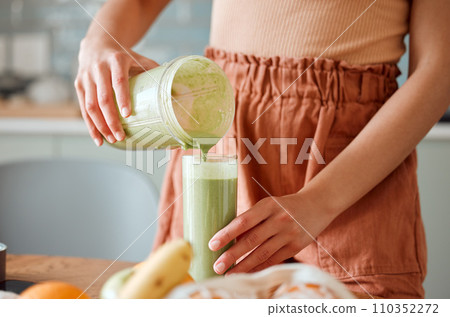 Woman pouring healthy smoothie in a glass from a blender jar on a counter for detox. Female making fresh green fruit juice in her kitchen with vegetables and consumables for a fit lifestyle. Woman pouring healthy smoothie in a glass from a blender jar on a counter for detox. Female making fresh green fruit juice in her kitchen with vegetables and consumables for a fit lifestyle. 110352272