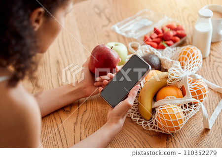 Colorful, healthy and nutritional organic fruit groceries on table beside woman holding phone and looking online for smoothie recipes. Young lady eating fresh organic apple for wellness vegan diet Colorful, healthy and nutritional organic fruit groceries on table beside woman holding phone and looking online for smoothie recipes. Young lady eating fresh organic apple for wellness vegan diet 110352279