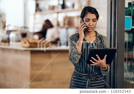 Female cafe business owner using phone talking, ordering and reading on tablet in her store. Serious businesswoman, entrepreneur or employee standing in a coffee shop preparing online grocery order. Female cafe business owner using phone talking, ordering and reading on tablet in her store. Serious businesswoman, entrepreneur or employee standing in a coffee shop preparing online grocery order. 110352543