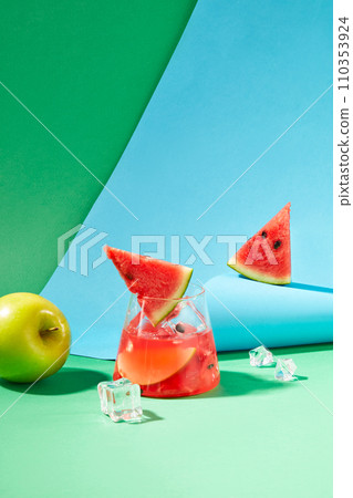 A glass of watermelon juice with ice, a green apple and a blue paper sheet on a green background. Supplement vitamins from fruits. Summer air with cooling water. A glass of watermelon juice with ice, a green apple and a blue paper sheet on a green background. Supplement vitamins from fruits. Summer air with cooling water. 110353924