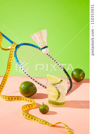 A glass of lemonade with juicy fresh lemons, a tape measure, a badminton racket and a shuttlecock on a pink surface with a green background. Healthy living concept. A glass of lemonade with juicy fresh lemons, a tape measure, a badminton racket and a shuttlecock on a pink surface with a green background. Healthy living concept. 110353931