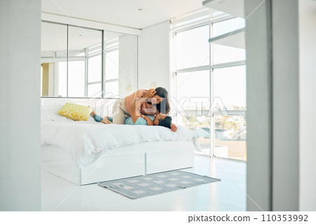 Happy playful young mixed race woman kneeling on top of her boyfriend in bed at home. Romantic couple bonding in their bedroom in the morning. Carefree boyfriend and girlfriend laughing while intimat 110353992