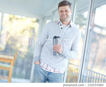 Portrait of man smiling and standing holding a flask. Attractive male leaning against a window inside his home with a travel coffee mug in his hand. Mature male drinking tea and enjoying free time 110355984