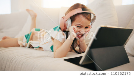 Happy little girl, tablet and headphones on sofa for elearning, entertainment or streaming at home. Female person, child or kid smile on technology in relax for movie, education or learning at house 110362175