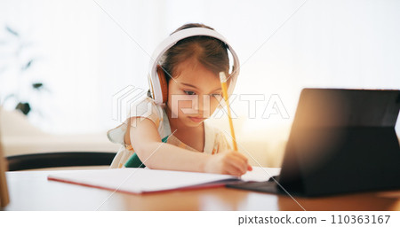 Girl, headphones and writing in book for elearning, education or study on desk at home. Female person, child or kid taking notes with tablet, headset or technology for virtual class at house 110363167