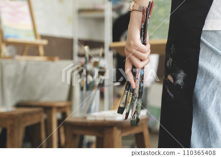 A close-up of the artist hands wearing an apron smeared with paint. Clutching many brushes and paintbrushes. A close-up of the artist hands wearing an apron smeared with paint. Clutching many brushes and paintbrushes. 110365408